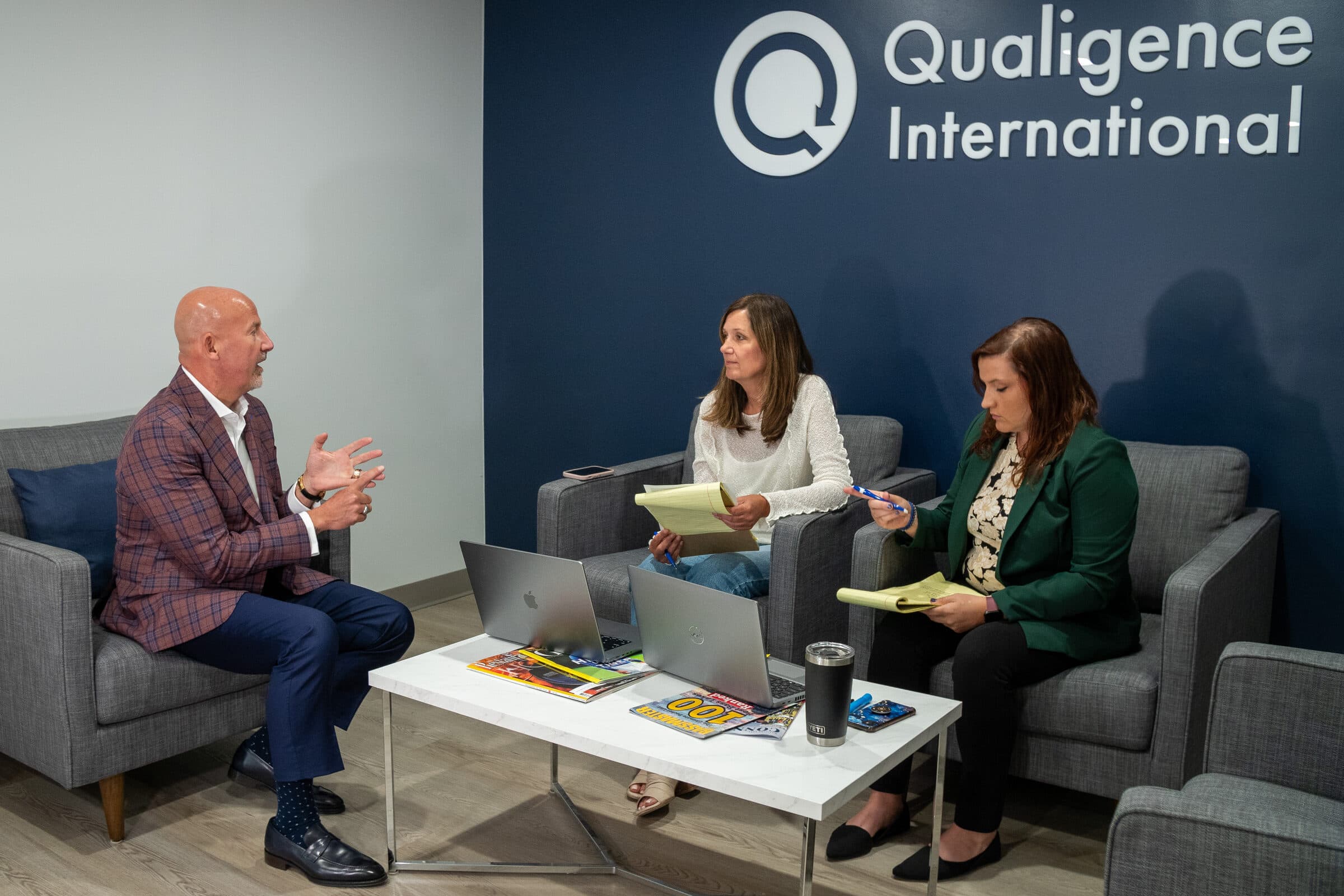 Steve Lowisz in conversation with two Qualigence team members beneath the Qualigence International logo wall